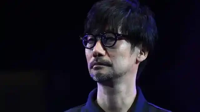A close-up portrait of a man with short black hair and glasses, wearing a dark shirt, looking thoughtfully to the side against a blurred dark background.
